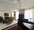 Elegant living room with a grand piano, fireplace, chandelier, and large windows with curtains. Features a mix of seating including a sofa, armchairs, and a dining table with chairs.