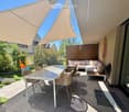 A modern outdoor patio with a large sunshade, a white dining table with four chairs, a sofa with cushions, and a small bench. The area is surrounded by greenery and has a view of the garden.