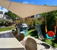 Outdoor patio with a table and chairs, a small playhouse, a slide, and a swing set, surrounded by greenery and a large shade sail.