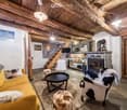 Cozy living room with exposed wooden beams, a yellow sofa, a black coffee table, a white armchair with a black and white cowhide throw, and a floor lamp. The room features a rustic wooden ceiling and a staircase leading to an upper level.