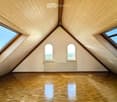Empty attic room with wooden slanted ceilings, wooden flooring, and multiple windows including skylights and arched windows, with a radiator below the windows.