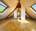 Bright, open attic space with wooden floors and slanted wooden ceilings. Large skylights and windows provide natural light, with a visible staircase leading to another level.