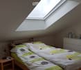Cozy attic bedroom with a skylight window, a double bed with a colorful duvet, a wooden nightstand with a fan and remote controls, a white dresser with a plant and a stuffed toy, and framed pictures on the walls.