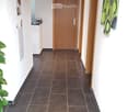 Clean hallway with dark tiled flooring, white walls, wooden doors, and exposed wooden beams on the ceiling. Features a small shelf with books and a plant on the left side.