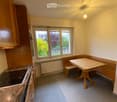 Compact kitchen with wooden cabinets, an induction stove, a small dining table with bench seating, and large windows overlooking a garden.