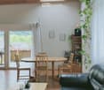 A cozy living room with a black leather sofa, wooden coffee table, dining table with chairs, and a shelf with books and a plant. The room features wooden walls, tiled flooring, and large windows with blinds.