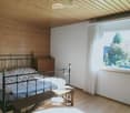 Cozy bedroom with wooden walls and ceiling, featuring a metal-framed bed, nightstand, lamp, and bench.