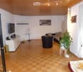 Living room with wooden flooring, white walls, and a slanted wooden ceiling. Features include a sofa, coffee table, armchair, potted plant, TV, cabinet, small table, and picture frame.