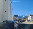 A multi-story building with a modern facade, parked cars in front, and a scenic mountain view in the background.