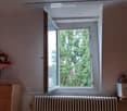 A small room with a large window showing greenery outside. The room features a wooden cabinet with a flower arrangement, a radiator below the window, and a small toy on a shelf.