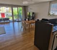 Open-plan living and dining area with a modern kitchen, large windows, and a balcony view. Features include a sofa, dining table, chairs, and a kitchen counter with a sink and dishwasher.