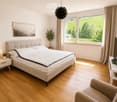 Modern bedroom with a large bed, nightstand, lamp, chair, screen, plant, and picture on the wall. Large windows offer a scenic view of a green landscape.