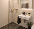 Modern bathroom with a glass-enclosed shower, a white sink with a mirror, and a toilet. The space features beige tiles, a dark grey floor, and decorative plants.