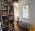 A cozy, well-lit room with wooden floors, a large bookshelf filled with books, a small grey chair, a yellow toy chair, a mirror with a decorative clock, and a small table with a backpack nearby.