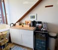 Compact kitchen with white cabinets, wooden countertop, coffee maker, refrigerator, and small cart. Features a slanted ceiling and a window with a view.