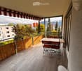 Balcony with tiled floor, wooden table, chairs, and a view of mountains and cityscape.