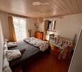 Cozy bedroom with a large bed, nightstand, wooden floor, and a small cradle. The room features large windows with curtains and a hanging pendant light.