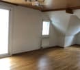 Empty room with wooden flooring, white walls, and a slanted wooden ceiling. Features include a large window, a smaller window, a radiator, and a chandelier light fixture.