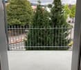 Balcony with metal railing, overlooking a green area with trees and a street.