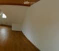 Empty room with wooden flooring, white walls, large window with glass door leading to a balcony, and visible ceiling beams.