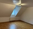 Empty attic room with wooden flooring, slanted wooden ceiling, single window, and a radiator. No furniture or equipment present.