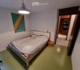 A small bedroom with a wooden bed, a pendant light fixture, a poster on the wall, and a small wooden stool near the bed. The room has light green flooring and white walls.