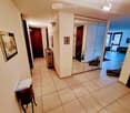 Hallway with tiled floor, white walls, several doors, and a small table with papers on it. A mirror and framed pictures are visible on the walls.