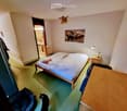 A small bedroom with a single bed, nightstand, wooden table, and a cat sitting on the floor. The room features a large mirror, a picture of elephants on the wall, and a small decorative angel figurine hanging from the ceiling.
