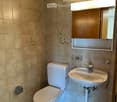Modern bathroom with beige tiled walls and floor, featuring a toilet, sink, mirror, and wooden cabinet with a built-in light fixture above it.