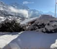 Snow-covered outdoor area with trees and mountains in the background, featuring a small wooden structure partially covered in snow.