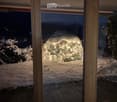 View from inside a house through large glass doors onto a snowy garden at night, with a snow-covered dome-shaped structure illuminated by lights.