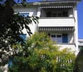 A multi-story residential building with white walls, balconies, and blue shutters on the windows. The building is partially obscured by green foliage in the foreground.