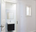 Modern bathroom with white walls, wooden flooring, a white sink with a black cabinet, and a mirror above it. The adjacent room has a small window and white walls.