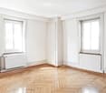 Empty room with white walls, wooden herringbone flooring, two windows, and radiators.