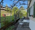 Balcony with metal frame, white table and chairs, potted plants, and a view of a garden and stone wall.