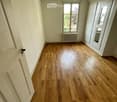 Empty room with wooden flooring, white walls, and large windows letting in natural light. Features a radiator and a hanging light fixture.