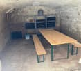Underground room with stone walls, a long wooden table with benches, a metal wine rack, and a metal pot on a shelf.