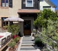 A charming garden path leads to the entrance of a house, flanked by potted plants and a small seating area with a white sofa and umbrella.
