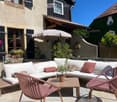 Outdoor patio with a white sectional sofa, pink cushions, a wooden coffee table, pink chairs, and a large umbrella. Potted plants and a stone house with shutters are visible in the background.