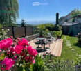 Well-maintained garden with a wooden deck, outdoor furniture, and colorful flowers, surrounded by greenery and a wooden fence.