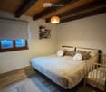 Cozy bedroom with wooden ceiling beams, a large bed with pillows and a blanket, a small wooden cradle, a window with a curtain, and a wall-mounted picture frame.