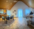 A modern, open-plan living and dining area with a slanted wooden ceiling, tiled floor, and large windows with curtains. The living area features a grey sectional sofa with throw pillows and a fur throw, a fireplace, and a dining table with four chairs. Th