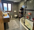 Modern bathroom with dark grey tiles, a large bathtub with a glass enclosure, a wooden sink cabinet, a toilet, and a shelf holding folded towels and toiletries.