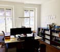 Office space with a large desk, dual monitors, chairs, shelves filled with books and files, and a fan. Natural light from large windows.