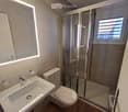 Modern bathroom with a white sink, toilet, glass-enclosed shower, and a small window above the shower. The walls are tiled, and the floor is wooden.