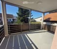 Balcony with metal railing, concrete floor, and large windows overlooking a residential area with trees and houses.