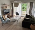 Modern living room with a grey sofa, glass coffee table, light blue armchair, TV stand, pendant lamp, and shelves with decorative items. Large windows with sheer curtains lead to a balcony.