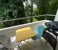 Balcony with a yellow table, two chairs, and a grill, surrounded by greenery.