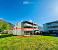 A modern building with a green exterior, surrounded by a well-maintained lawn and a tree. The building has multiple floors with balconies and windows. In the background, there are mountains and another building.