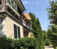 Two-story house with beige walls, dark green shutters, and a shingled roof. Features a balcony with railings and Swiss flags, surrounded by lush greenery and trees.